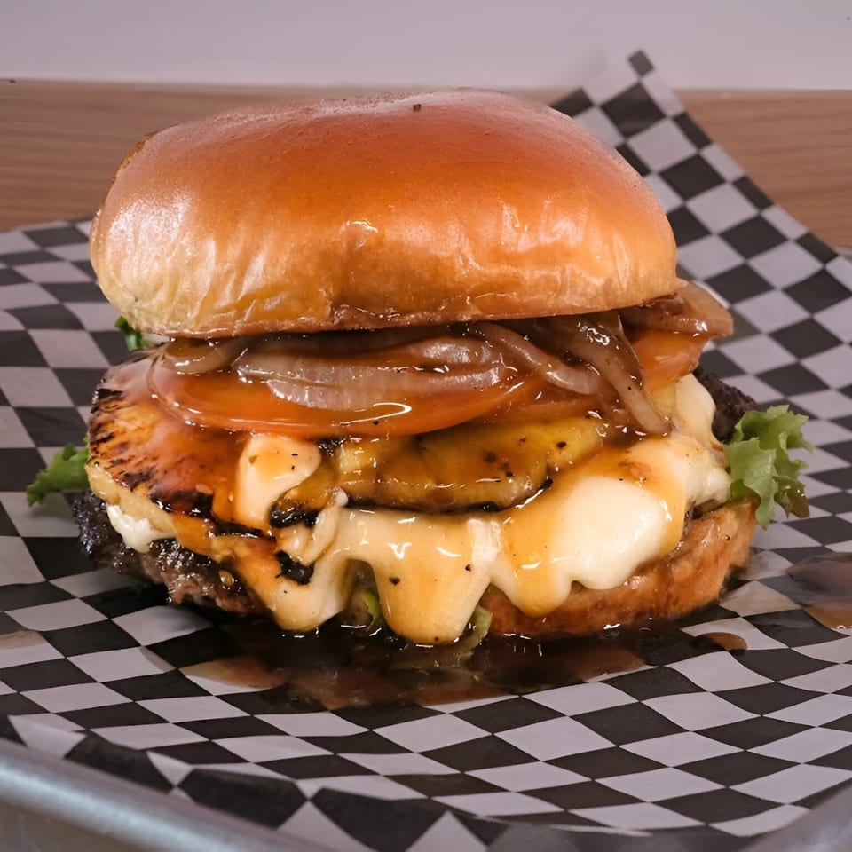 Made Burgers' burger with melted cheese, grilled pineapple, caramelized onions, and sauce on checkered paper.