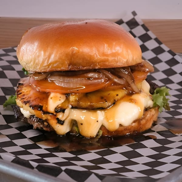 A Made Burgers burger with grilled pineapple, melted cheese, caramelized onions, and sauce on checkered paper.