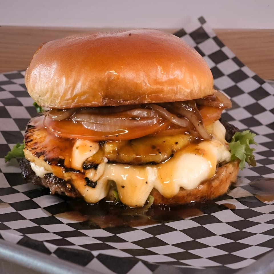 A Made Burgers burger with grilled pineapple, melted cheese, caramelized onions, and sauce on checkered paper.