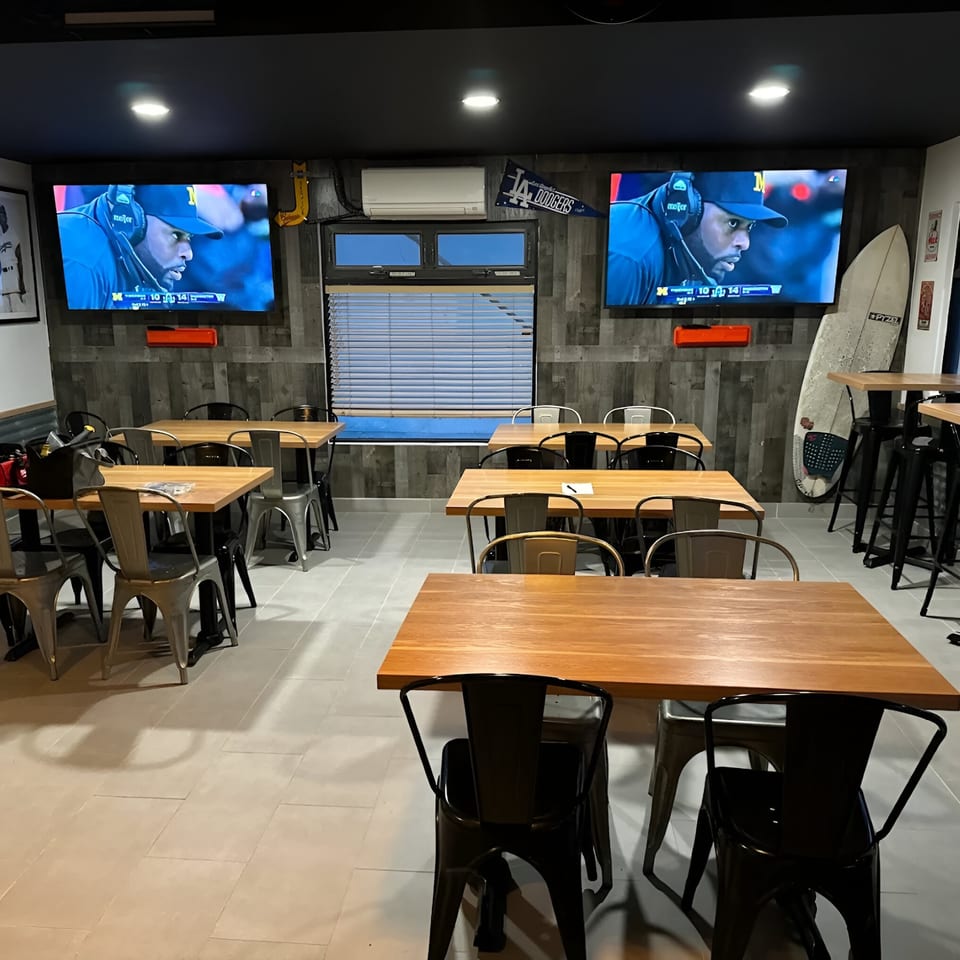 Interior of Made Burgers with wooden tables, metal chairs, and two TVs displaying a football game.