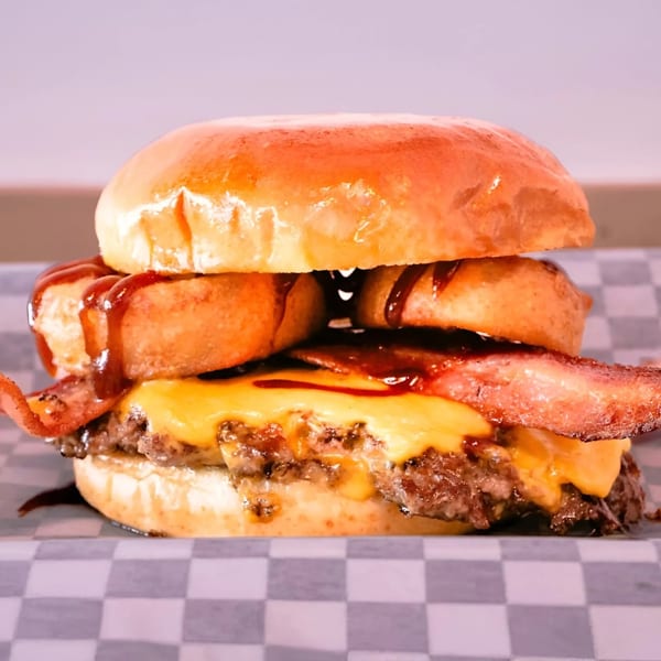 Made Burgers' bacon cheeseburger with onion rings and BBQ sauce on a checkered paper.